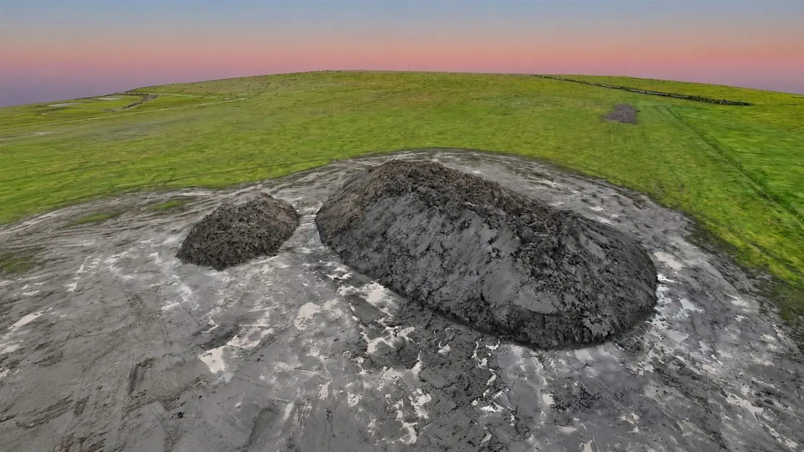 Measurable 3D surface model of two soil stockpiles on a green paddock at dusk, reconstructed from overlapping drone photographs